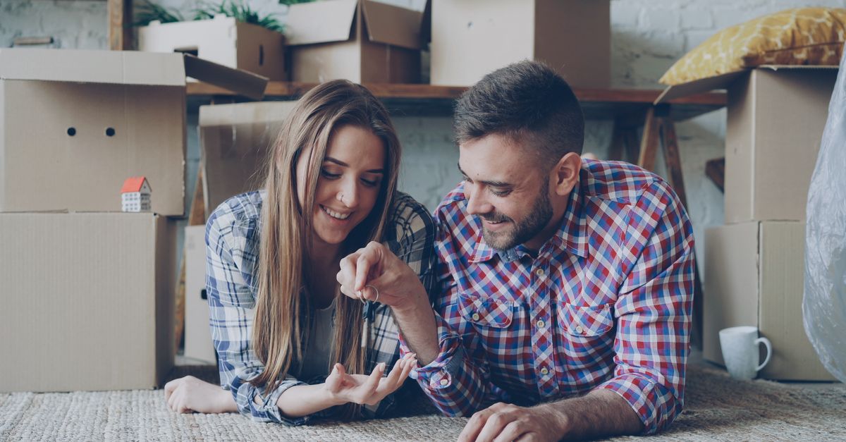 Joyful couple laying on floor surrounded by moving boxes, holding keys to their new home.