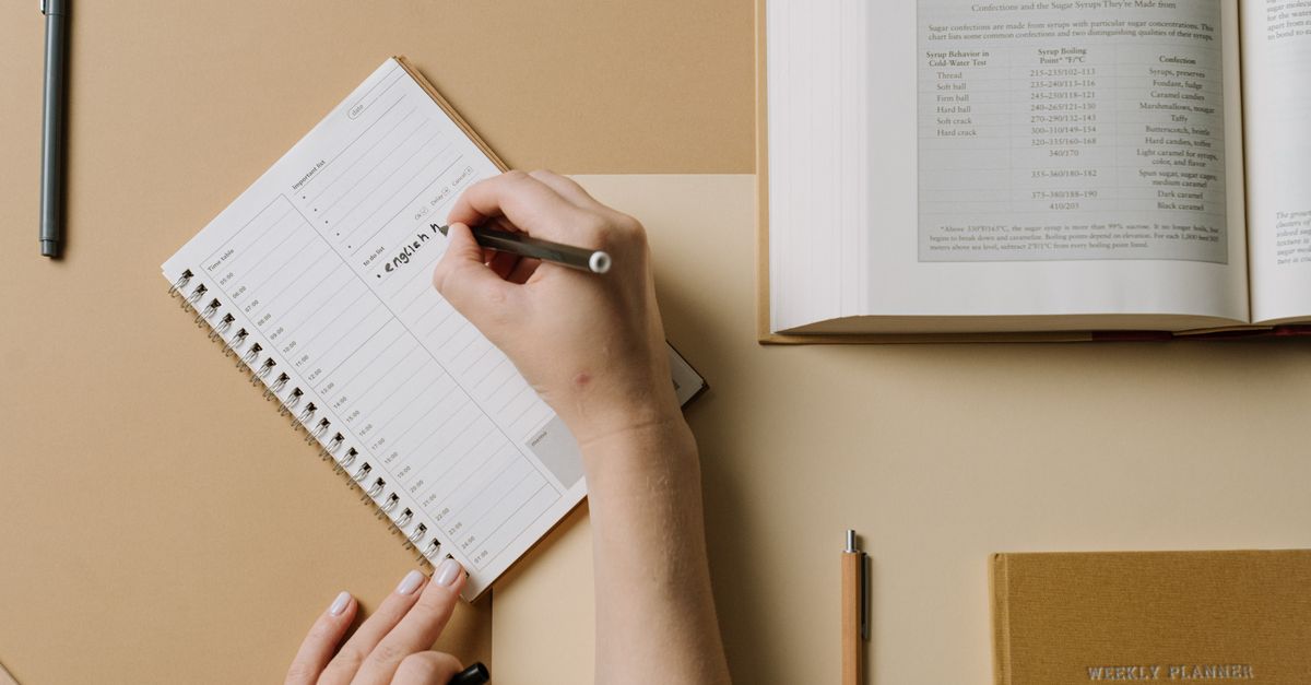 A person's hands writing in a planner at a desk with an open book nearby, conveying an organized workspace.